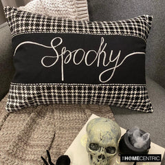 A Halloween throw pillow featuring 'Spooky' in script with black and white houndstooth borders, complemented by a skull and spider for a spooky touch, set on a grey chair with textured grey throw.