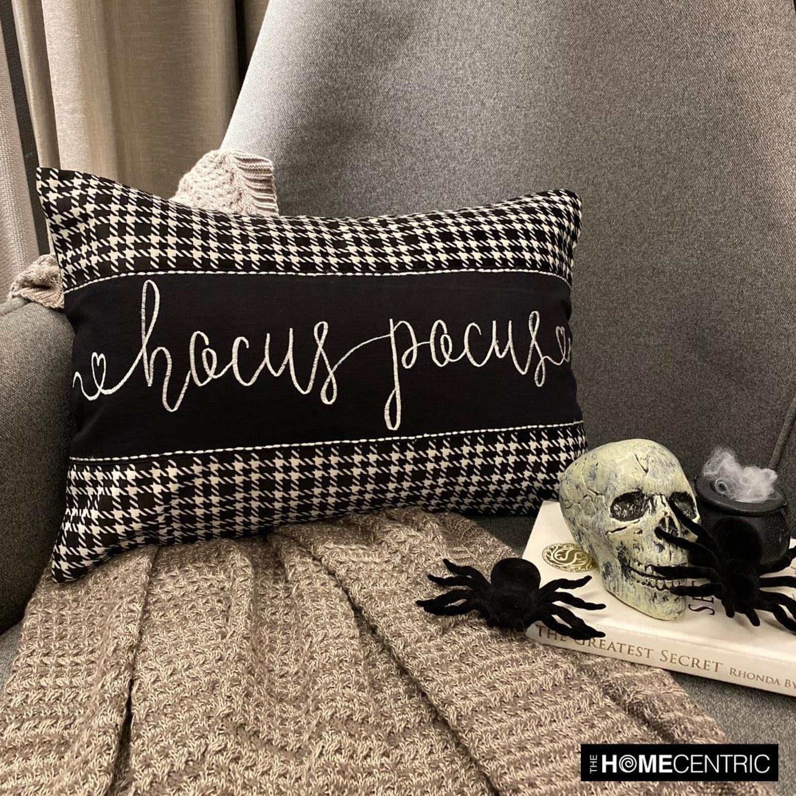 Hocus Pocus themed cotton, houndstooth patterned pillow cover with cursive white lettering, set against a gray chair and a gray throw, surrounded by Halloween-themed decor like a skull and spiders.