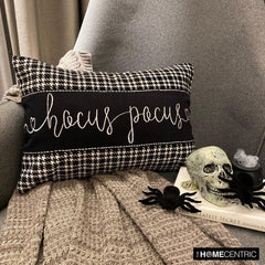 Hocus Pocus themed cotton, houndstooth patterned pillow cover with cursive white lettering, set against a gray chair and a gray throw, surrounded by Halloween-themed decor like a skull and spiders.