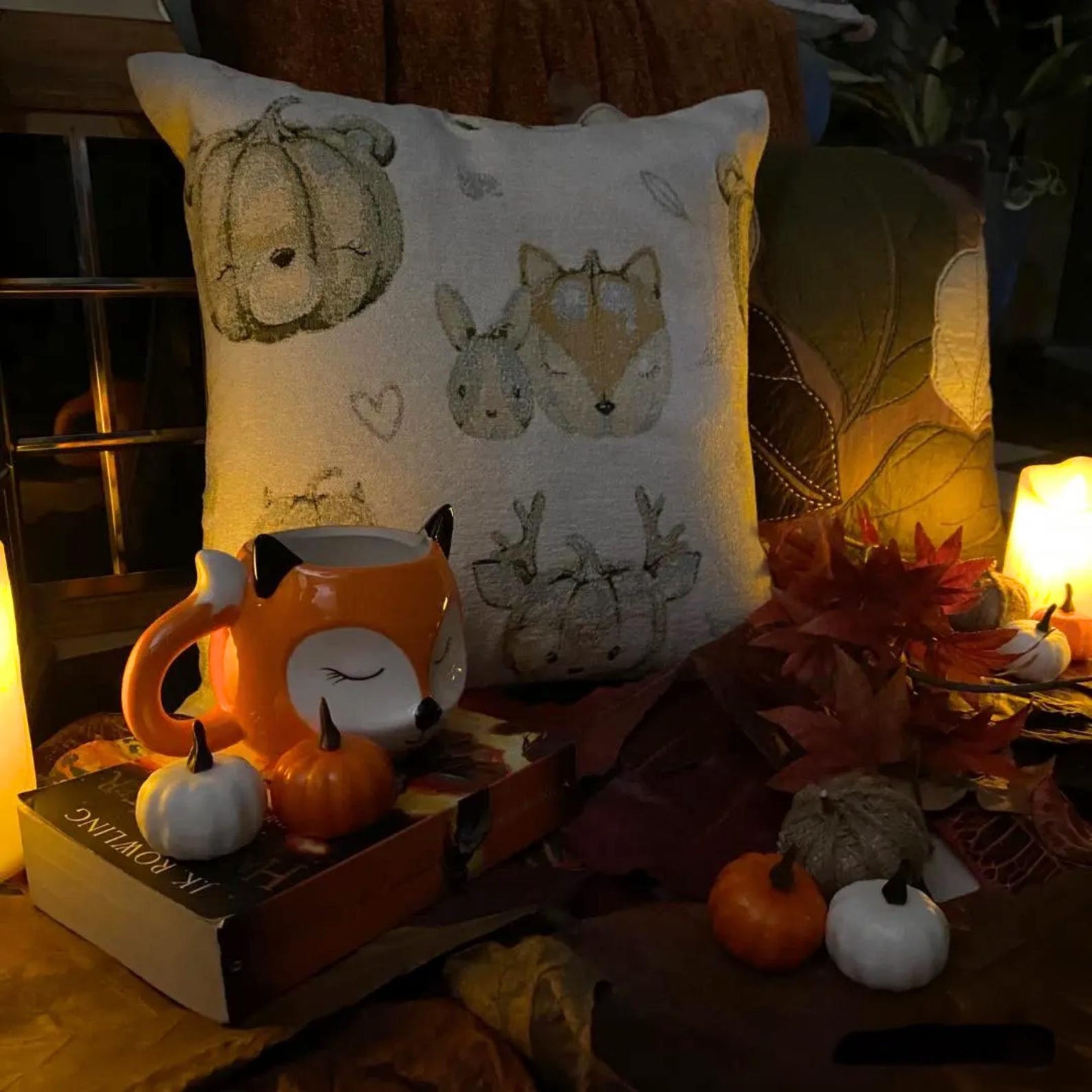 Fall Halloween home decor pillow cover, A woven jacquard throw pillow cover with pumpkin fox design and subtle fall colors for Autumn Decor set in a cozy fall evening setting with books, hot chocolate mug and small decorative pumpkins and maple leaves, elevated with candles and pillow covers.  Handcrafted pillow cover from The HomeCentric's Fall Collection.
