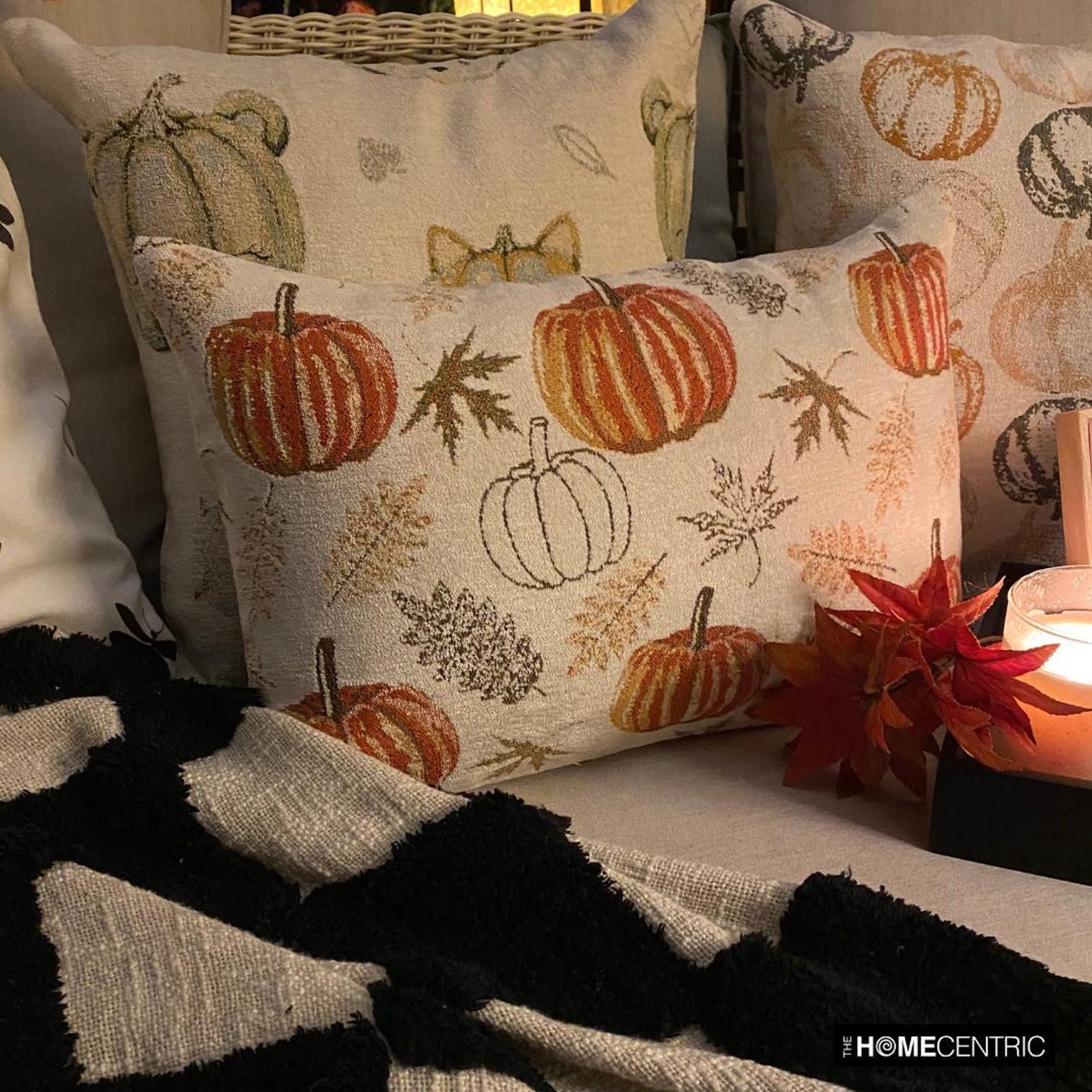 A cozy fall setup by The HomeCentric highlighting their Autumn Decor collection with five pillow covers showcasing the fall season, set on a couch decorated with books, hot chocolate mug, candles, pumpkins and maple leaves. 