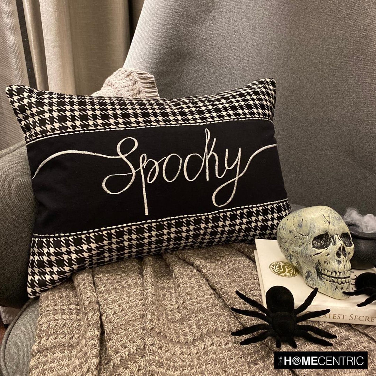 Stylish Halloween-themed throw pillow with the word 'Spooky' in white cursive on a black fabric with houndstooth pattern accents, paired with a skull, spider, a witch pot with white flames on a book, placed on a grey chair with a textured grey throw.