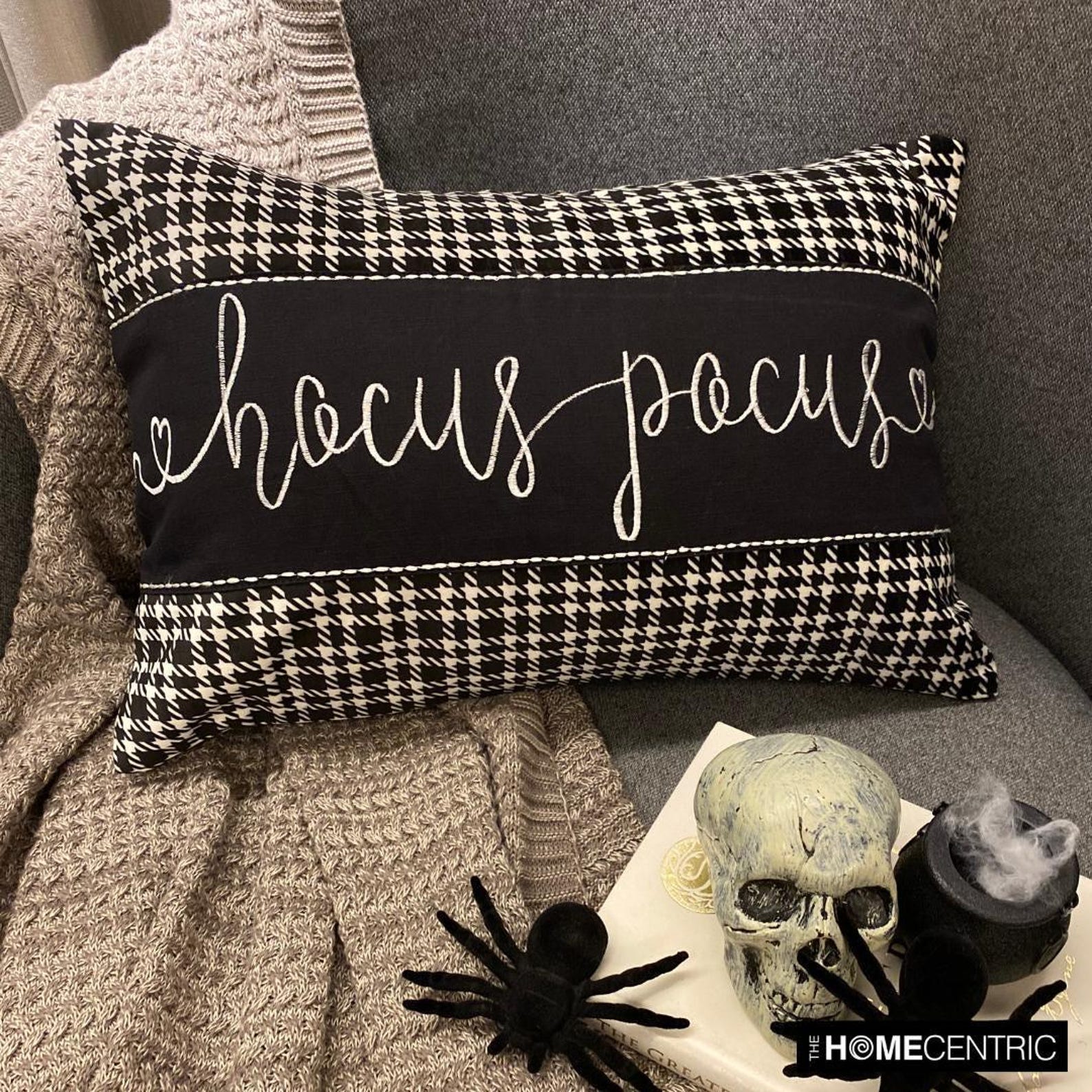 Decorative oblong pillow cover with 'Hocus Pocus' text in cursive on a black and white cotton houndstooth fabric, placed on a cozy gray throw with a skull and spider decorations for Halloween Decor.