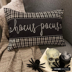 Decorative oblong pillow cover with 'Hocus Pocus' text in cursive on a black and white cotton houndstooth fabric, placed on a cozy gray throw with a skull and spider decorations for Halloween Decor.