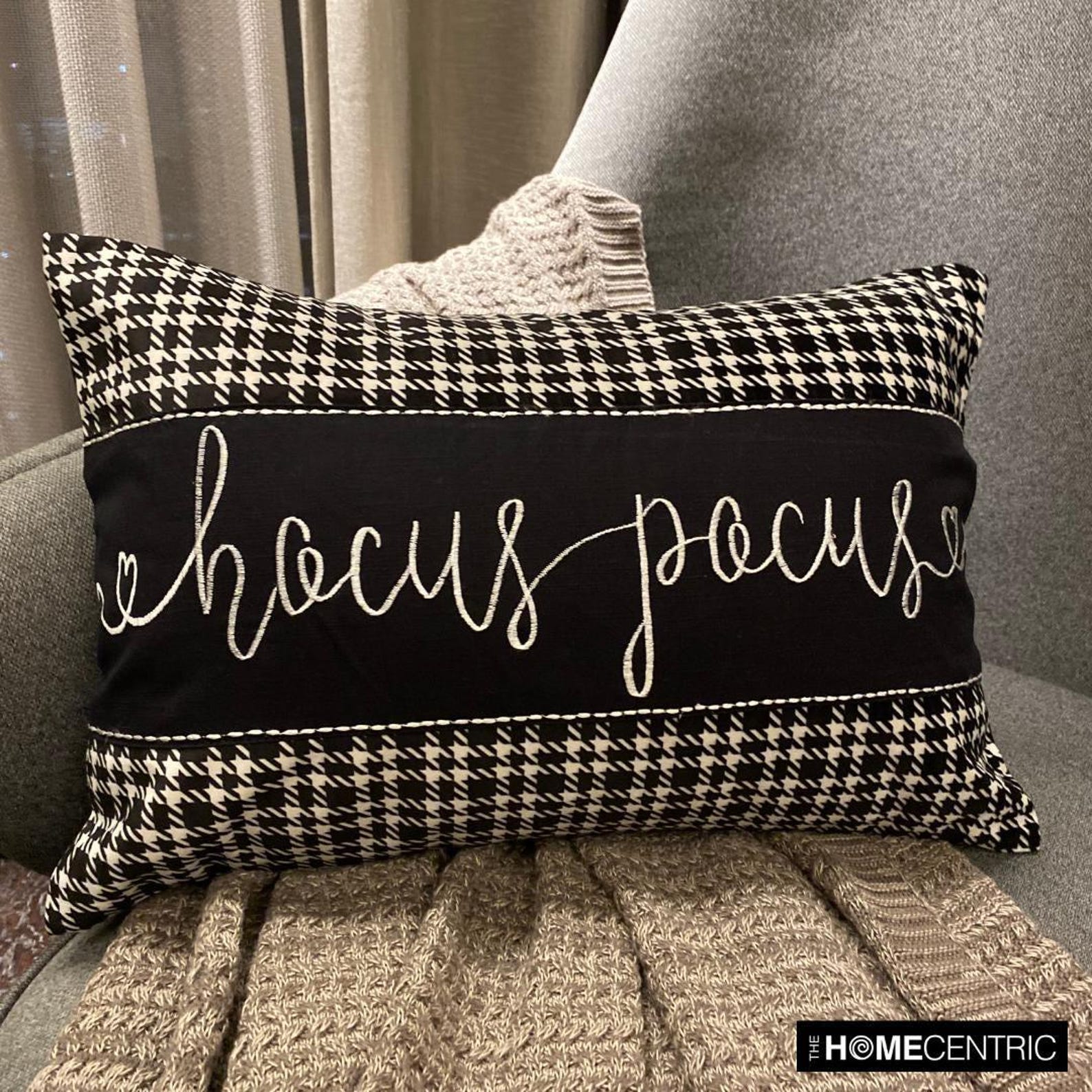 Halloween movie-themed pillow cover with 'Hocus Pocus' text in white cursive, featuring a houndstooth pattern, placed with skull and spider decorations on a cozy gray throw on a gray chair.