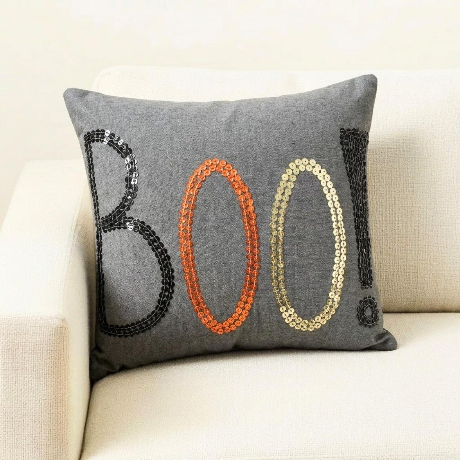 Halloween-themed throw pillow cover with the word 'BOO!' in black, orange, and gold sequins, placed on a black and white monochrome throw against a wooden background. 
