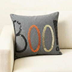 Halloween-themed throw pillow cover with the word 'BOO!' in black, orange, and gold sequins, placed on a black and white monochrome throw against a wooden background. 