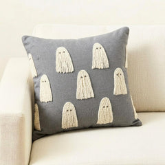 Skeleton holding a grey spooky ghost pillow cover surrounded by Halloween decor such as a skull and a witch pot placed on a book.