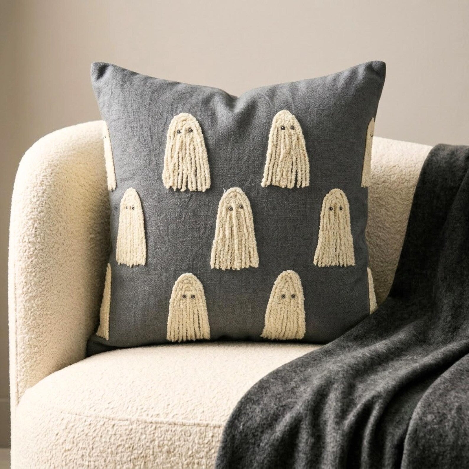 Halloween themed skeleton holding a grey decorative embroidered throw pillow cover with cute ghost design placed on a black chair covered with spider web, decorated with a skull and a witch pot kept on a black book to create a Halloween look. 