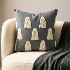 Halloween themed skeleton holding a grey decorative embroidered throw pillow cover with cute ghost design placed on a black chair covered with spider web, decorated with a skull and a witch pot kept on a black book to create a Halloween look. 