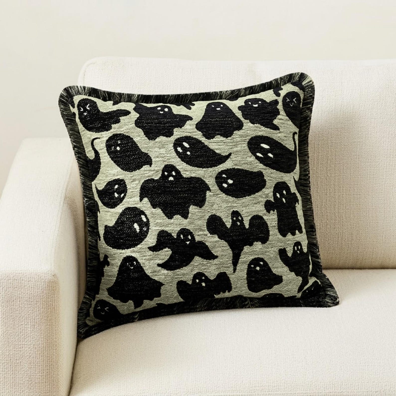 Halloween-themed pillow cover featuring ghost print on neutral fabric, set on a black chair with a checkered black & white throw against a brown wood background.