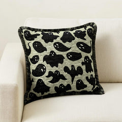 Halloween-themed pillow cover featuring ghost print on neutral fabric, set on a black chair with a checkered black & white throw against a brown wood background.