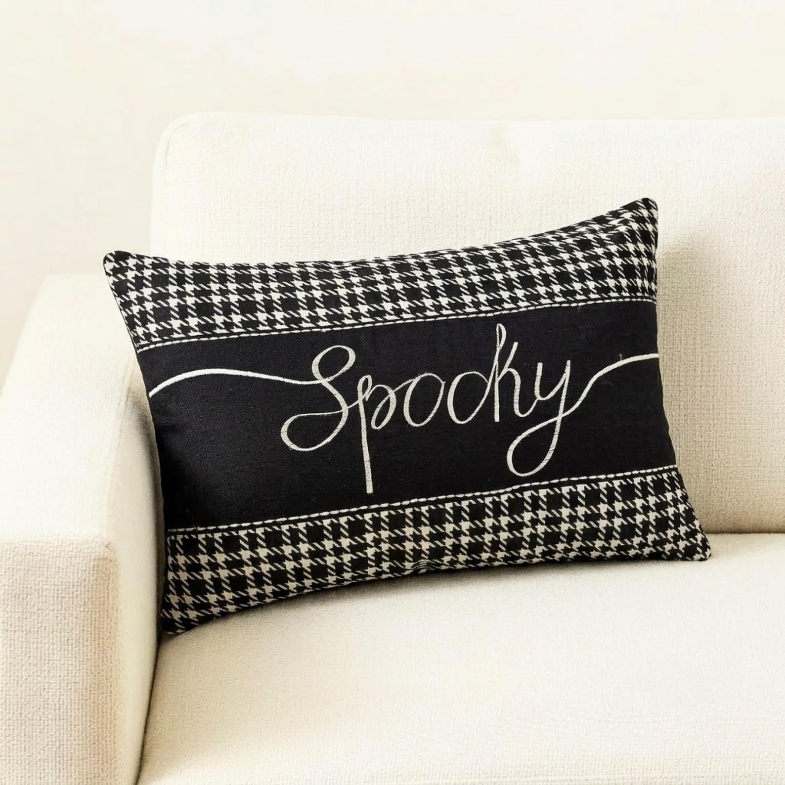 Halloween-inspired throw pillow with the word 'Spooky' written in a white, elegant script, set against a black and white houndstooth pattern, perfect for Halloween home decor, set on a grey textured throw on a gray chair.
