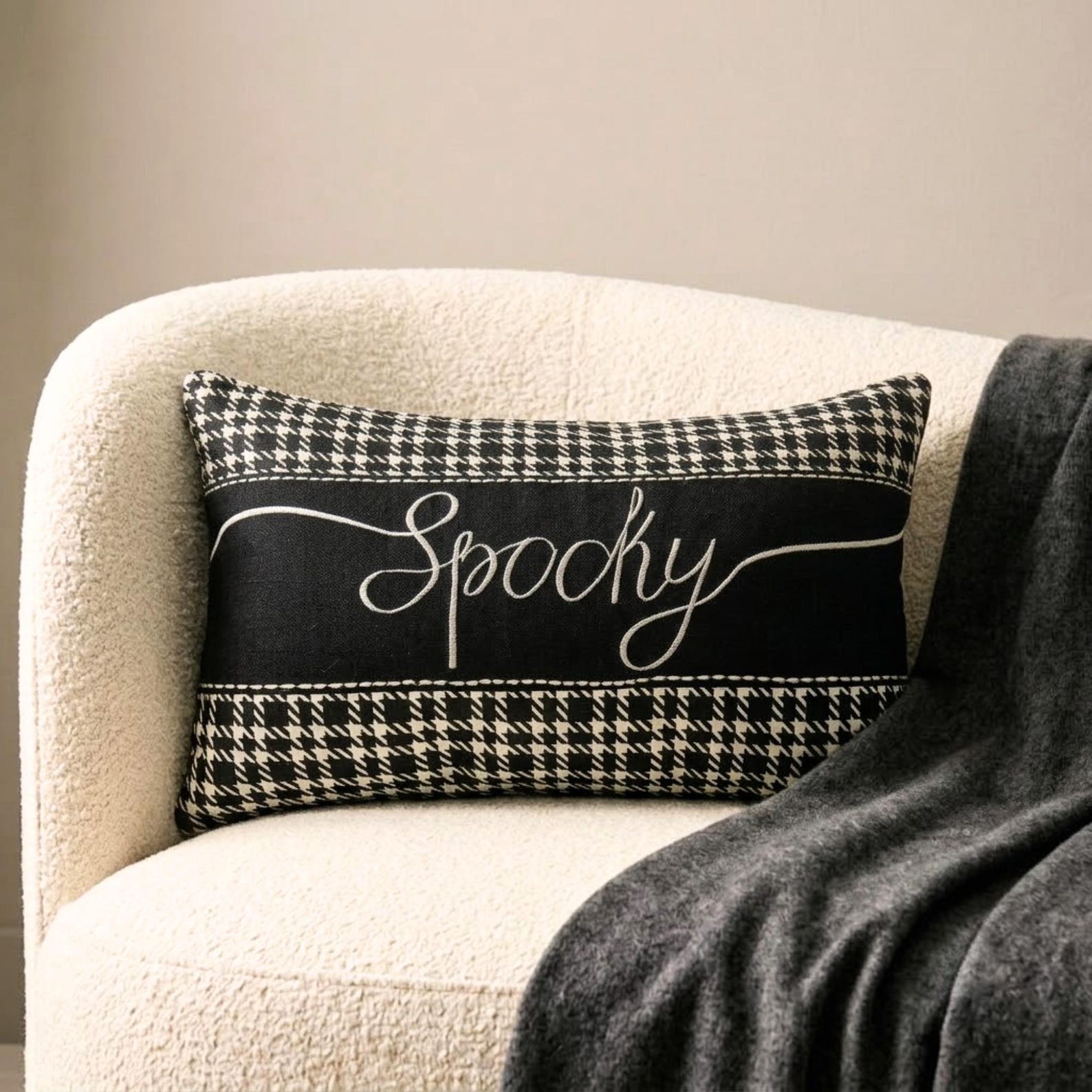 Halloween-inspired throw pillow with the word 'Spooky' written in a white, elegant script, set against a black and white houndstooth pattern, perfect for Halloween home decor. Set on a grey chair with a textured grey throw, surrounded by Halloween decor such as a skull, witch pot with white smoke, and a spider.