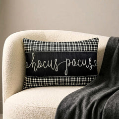 Black and white applique houndstooth lumbar pillow cover with embroidered 'Hocus Pocus' text on the center in cursive, placed on a gray couch and a gray throw with a skull and book in the foreground.
