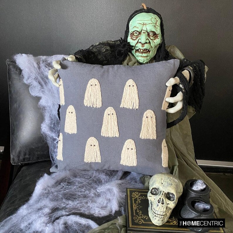 Skeleton holding a grey spooky ghost pillow cover surrounded by Halloween decor such as a skull and a witch pot placed on a book.
